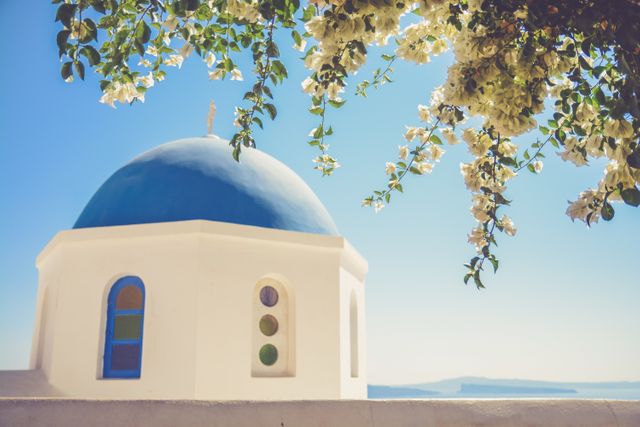 Iconic blue-domed church in Santorini, Greece, with flowering vines against a clear sky and serene sea background. Perfect for travel blogs, Mediterranean travel promotions, cultural context, and vacation planning. Ideal for introducing Greek architectural beauty and tourism projects.