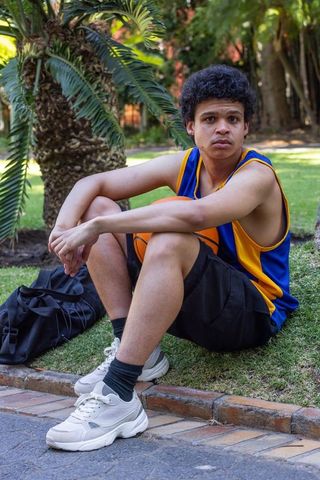 Young african man resting with basketball on grass in park wearing blue yellow jersey