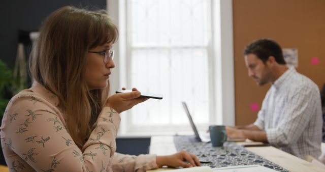 Young professionals are focusing on their respective tasks in a contemporary office environment. The female on the left side is holding a phone near her mouth, probably using a voice assistant, while the male in the background is working on a laptop. This image could be used for conveying themes of modern workspaces, technology use in professional settings, coworking dynamics, and communication in business.