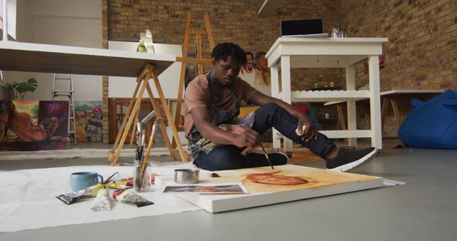The scene depicts a young male artist engaged in painting in a spacious, modern loft studio. Surrounding him are various art supplies including paint tubes, brushes, and an easel, emphasizing a creative and industrious atmosphere. This image can be used in articles or blogs about modern art, creative workspaces, and artist profiles, as well as in advertisements for art supplies or courses.