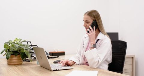 Female doctor consulting by phone while typing on laptop at minimalist clinic desk