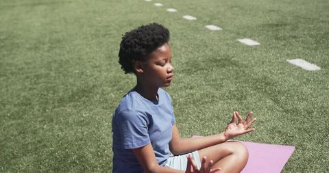 African american teen meditating on pink yoga mat on sports field in sunlight