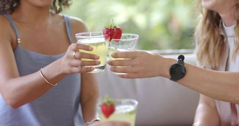 Diverse women clinking strawberry-garnished drinks on sunlit patio sofa showing smartwatch