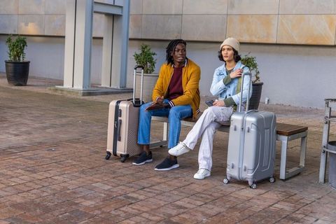 Young travelers sitting on bench waiting with luggage in modern urban plaza, diverse pair