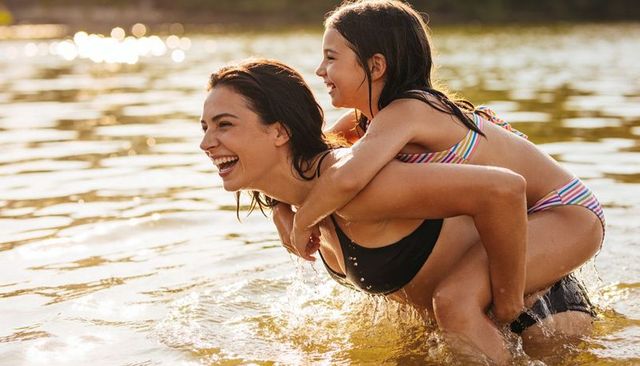 Mother giving daughter piggyback ride in lake at sunset smiling family summer outdoor fun