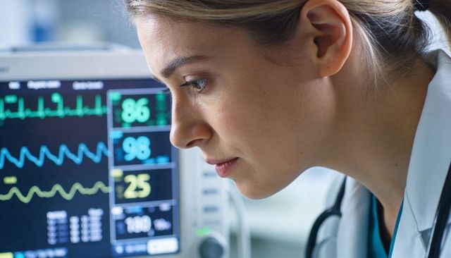 Clinician monitoring ecg and vital signs on bedside monitor wearing lab coat and teal scrubs