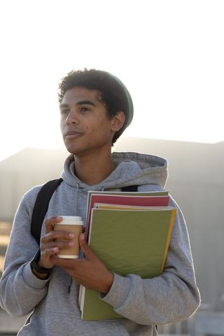 Thoughtful teen student holding coffee and colorful folders on urban campus