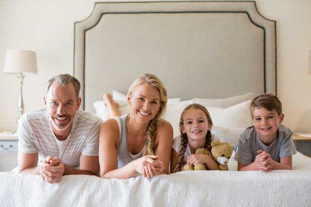 This image shows a happy family of four relaxing on a bed in a cozy bedroom. The parents and children are smiling, creating a warm and loving atmosphere. Ideal for use in family-oriented advertisements, parenting blogs, lifestyle magazines, and home decor promotions.