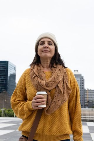 Confident middle-aged woman holding coffee in city plaza wearing mustard sweater and scarf