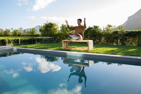 Asian man meditating on wooden bench by pool with mountain reflection, sunlit garden