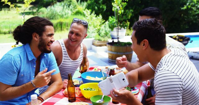 Four friends are enjoying a fun picnic in a backyard with green scenery in the background. They are seated at a red and white checkered table and appear to be eating and drinking. One of them is taking a selfie while others are laughing and engaging in conversation. This image is perfect for illustrating social gatherings, friendship, summer activities, and outdoor fun.