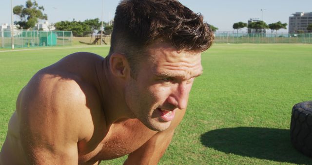 Image depicts a shirtless man who appears to be catching his breath in an outdoor grassy area. His posture and facial expression suggest intense physical exertion, likely from a rigorous workout. Use this image for fitness, sports, outdoor exercise promotions, health and wellness articles, or athletic training programs.