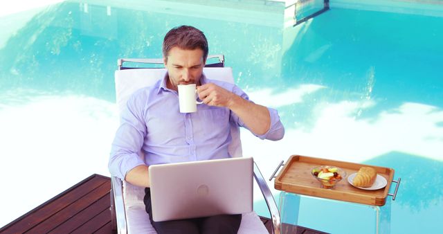 A businessman in a dress shirt enjoying a coffee while working on his laptop by the pool. A wooden tray with fruit and pastries is beside him. This is ideal for concepts of remote work, professional lifestyle, work-life balance, and leisure.