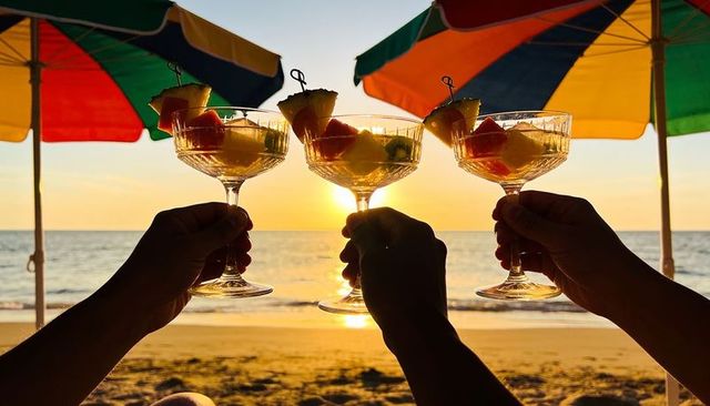 Toasting with tropical coupe cocktails at golden hour beach sunset under rainbow umbrellas