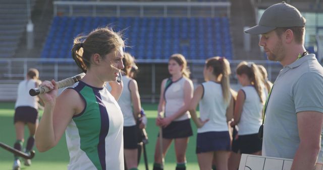 Female field hockey team practicing together on sports field while coach gives instructions. Players wearing uniforms holding field hockey sticks, engaging in team discussions and strategies. Perfect for sports education materials, team building, and promotional content for sports organizations.