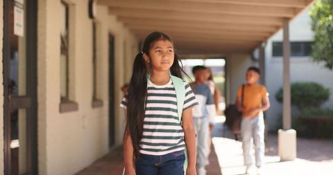 Asian teenage girl walking sunlit school corridor carrying backpack and looking at windows