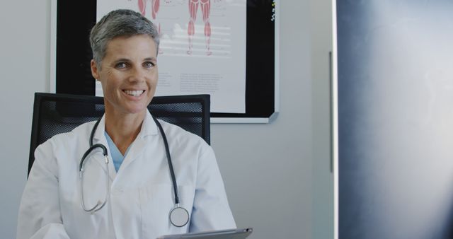 Mature female doctor smiling while holding a digital tablet in her medical office. Includes medical posters in the background and she is seated behind a desk. This is perfect for use in healthcare marketing materials, medical websites, and promoting patient consultations.