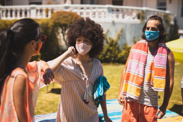 Diverse group of friends wearing face masks greeting each other with an elbow bump at an outdoor pool party. They are enjoying a sunny day while maintaining social distancing measures. This image can be used for promoting safe social interactions during the pandemic, summer activities, and health awareness campaigns.