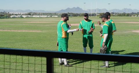 Diverse male baseball team huddling on field wearing green yellow uniforms planning play