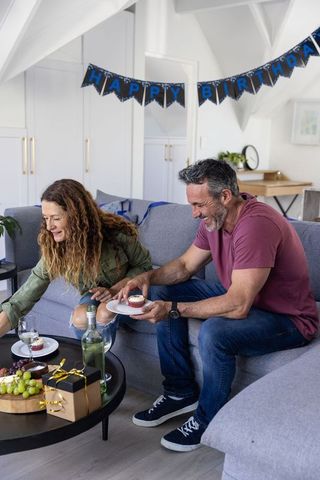 Middle-aged couple celebrating birthday on cozy sofa sharing cupcakes and wine