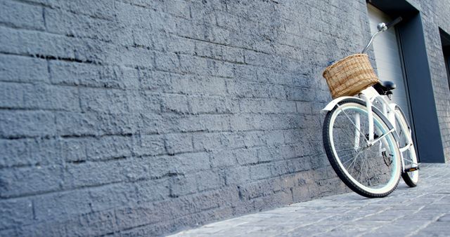 A vintage white bicycle with a wicker basket leans against a textured gray brick wall, with copy space. Its classic design adds a nostalgic touch to the modern urban environment.