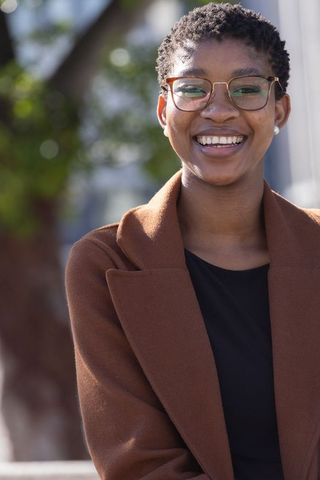 Smiling black woman wearing brown coat and glasses, urban headshot for branding