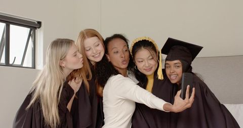 Multicultural female graduates taking joyful selfie on bed celebrating graduation success