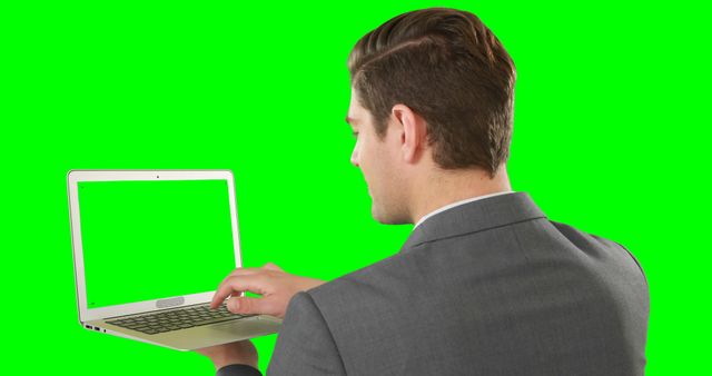 A businessman in a suit works on a laptop with a green screen background. Ideal for business, technology, remote work, digital marketing or corporate imagery. Can be used to illustrate business presentations or advertising campaigns requiring a professional setting.