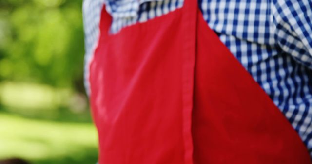A person wearing a red apron over a blue and white checkered shirt stands outdoors, with copy space. The apron suggests the individual could be engaged in outdoor cooking or gardening activities.