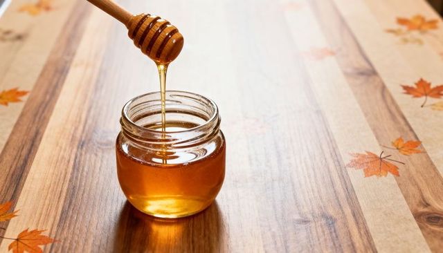 Dripping honey dipper releasing golden honey into glass jar on autumn wooden table