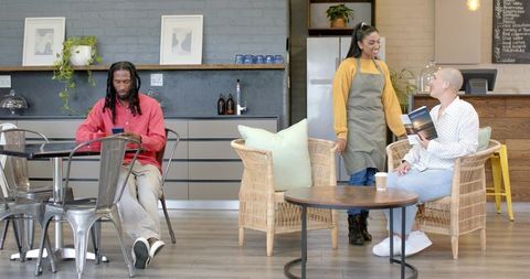Diverse barista chatting with customers in modern cozy cafe lounge with wicker seating