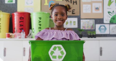 Smiling Schoolgirl Holding Green Recycling Bin in Classroom