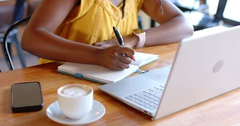 Woman writing notes in notebook while working on laptop in coffeehouse with cappuccino