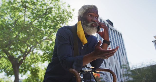 African american senior man wearing face mask talking on smartphone while leaning on his bicycle on the road. hygiene and social distancing during coronavirus covid-19 pandemic.