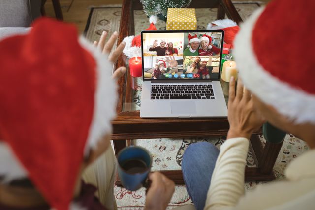 A diverse couple wearing Santa hats are engaged in a video call with their Caucasian friends, who are also in festive attire. The scene has Christmas decorations and gifts, suggesting a happy and connected holiday season despite physical distances. This image is perfect for illustrating technology's role in bringing together people during holidays, festive greetings, virtual family gatherings, and holiday marketing materials.