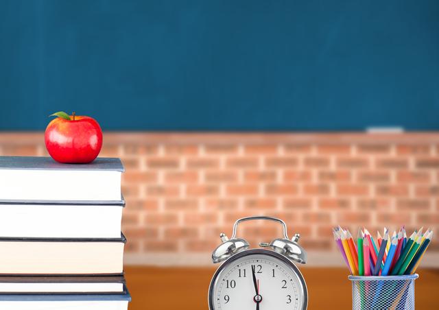 An image of an apple placed on a stack of books next to an alarm clock and colorful pencils in a metal holder with a classroom background. Perfect for representing education, teaching, learning environments, back-to-school concepts, and academic settings.