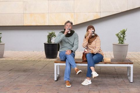 Middle-aged couple sipping coffee and eating apple while relaxing on urban bench in courtyard
