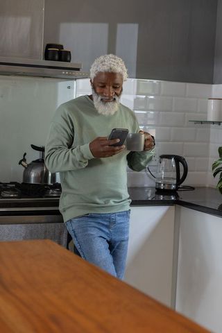 Senior african american man leaning at modern kitchen counter using phone and holding mug