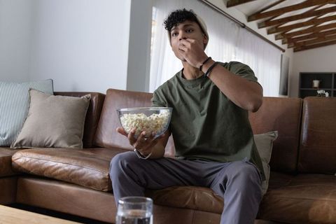 African american man eating popcorn on leather couch watching movie at home