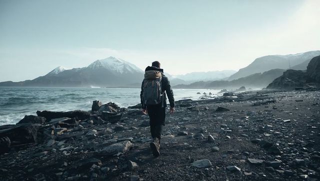 Solitary hiker walking rocky coastal shore toward snowcapped mountains under pale sky