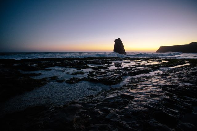 The image captures a breathtaking sunset over a rocky coastal shoreline, with waves crashing against the rocks and a prominent rock formation rising from the water. The twilight sky transitions from yellow near the horizon to deep indigo as it meets the night. Ideal for use in travel magazines, nature-themed websites, and meditation or relaxation apps, conveying a sense of peace and natural beauty.