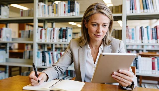 Focused professional woman using tablet and taking notes in library with bookshelves