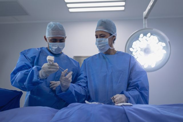 Two surgeons in blue scrubs and surgical masks perform a procedure in a sterile hospital operation theater. Bright surgical lights illuminate the scene. Suitable for medical, healthcare, hospital, and teamwork related concepts.
