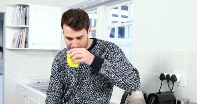 This photo shows a young man in a gray sweater drinking coffee in a bright modern kitchen. The man appears to be relaxed and enjoying his morning drink. Ideal for use in lifestyle blogs, coffee-related promotions, advertising casual morning routines, or illustrating a relaxed home environment.