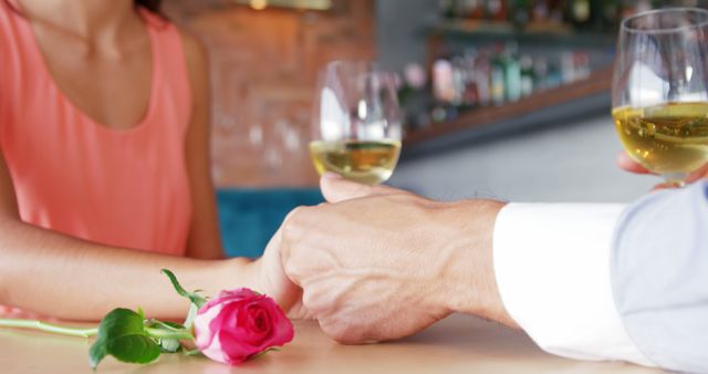 Couple enjoying a romantic evening at a cozy wine bar. Two people are toasting with glasses of white wine while holding hands, a single rose placed on the table signifying romance. Ideal for use in advertisements, articles, or promotions related to romance, dating, relationships, or special celebrations.