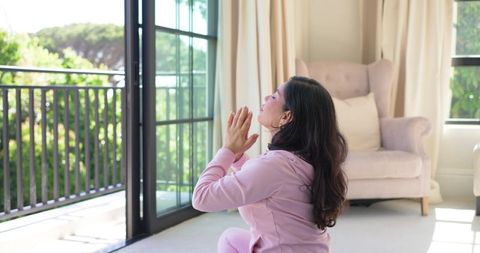 Woman practicing mindful meditation near sunlit balcony in cozy pink loungewear