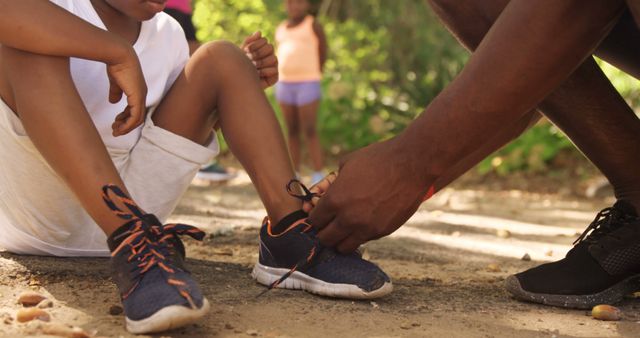 This scene shows a parent kneeling and tying the shoe of a child in a nature park. The image captures the essence of family bonding and care, perfect for use in family-focused advertisements, parenting blogs, or promotions for outdoor activities. The shoes, highlighted in the action, also make this image suitable for footwear brands. The lush greenery sets a serene backdrop, adding to the warmth of the moment.