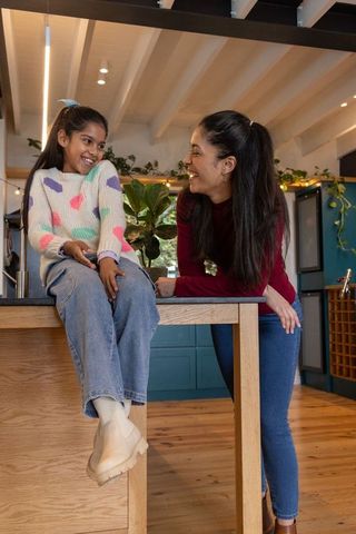 Indian mother and daughter laughing and chatting on kitchen island in cozy modern blue kitchen