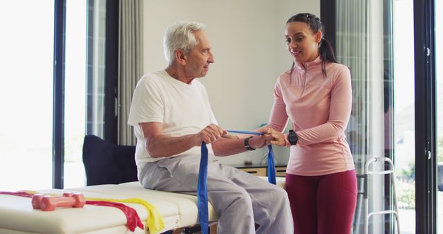 Senior man participating in a rehabilitation session with a young female therapist. They are using an exercise band for strength training. Setting is a bright, modern room with fitness equipment visible. Useful for illustrating healthcare, elder care, physical therapy, or active aging.