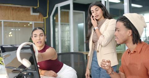 Young diverse team collaborating at monitor, pointing to screen, colleague talking on phone